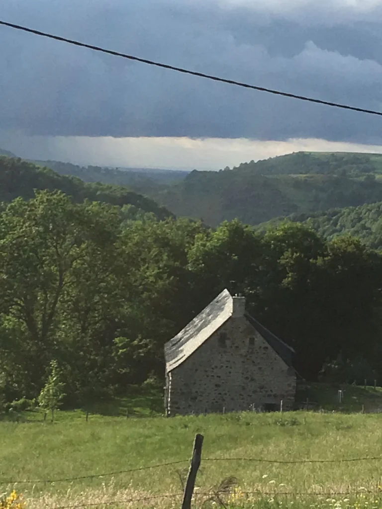 La Grange de L’Abille aus der Ferne, auf ihrer Wiese, mit dem bewaldeten Tal und bewölktem Himmel