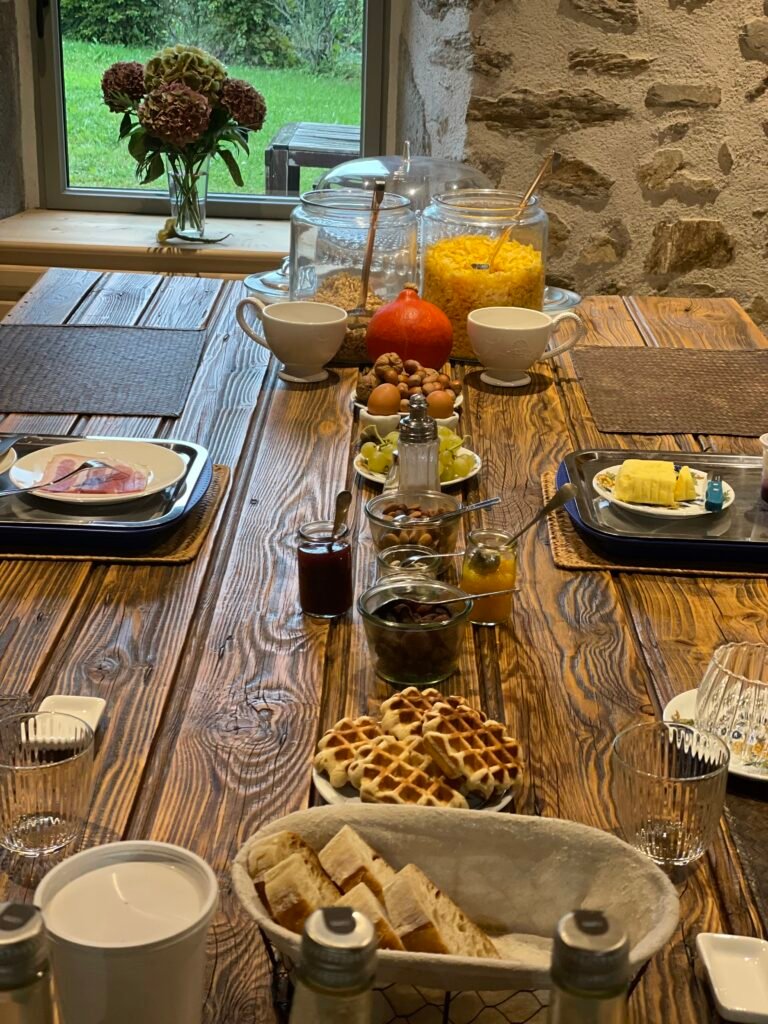 Table du petit-déjeuner : pain, gaufres, jus d’orange, confitures et fleurs devant une fenêtre sur le jardin