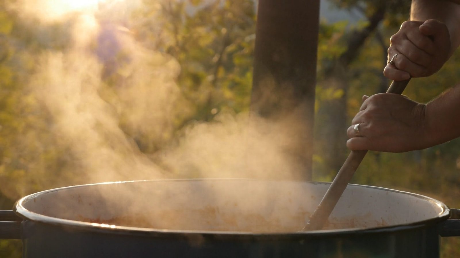 Des mains remuent une grande marmite qui fume, dans la lumière dorée du soir